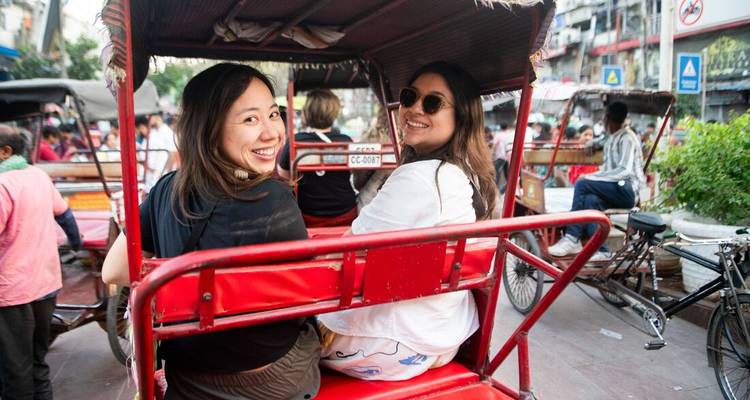 Two women smile from the back seat of a red cycle rickshaw navigating city traffic.