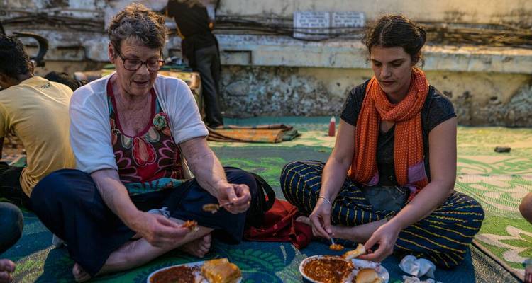 Travellers sit cross-legged on a mat outdoors sharing a simple street meal.