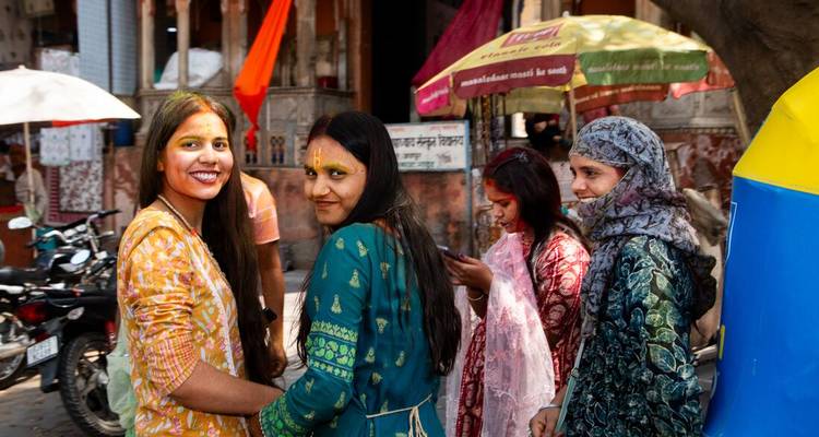 Young women with Holi colours on their faces smile in a lively street scene.