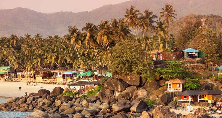 Rocky Goa coastline with colourful beach huts and leaning palms at sunset.