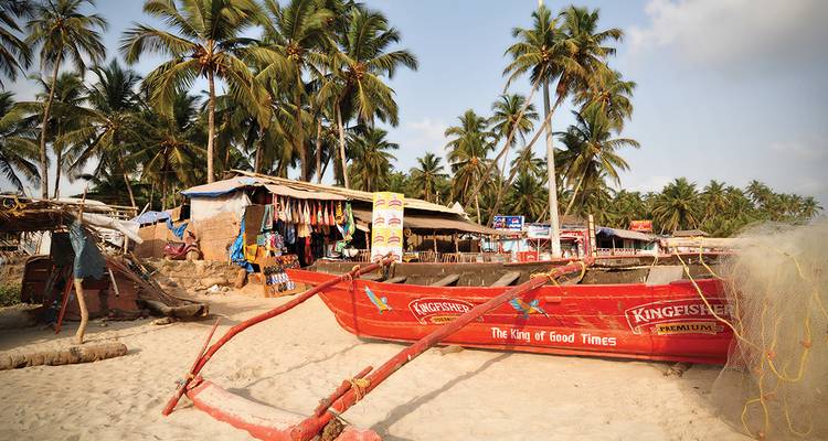 Red fishing boat and rustic beach shacks beneath swaying palms on a sandy Goan shore.