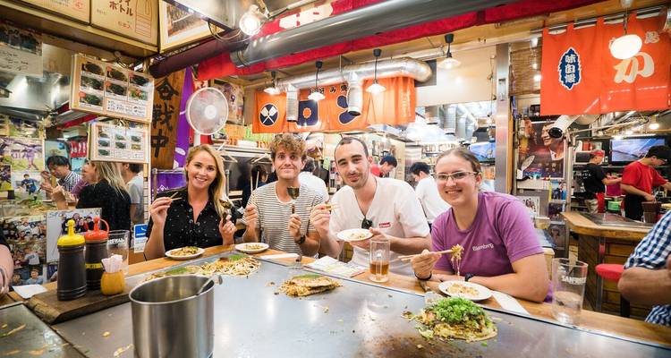Les voyageurs sourient autour d'un comptoir teppan garni d'okonomiyaki et de boissons dans un établissement de restauration animé.
