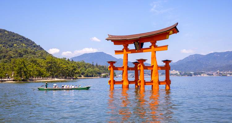 Le portique torii vermillon emblématique du sanctuaire d'Itsukushima s'élève des eaux bleu calme avec des montagnes en arrière-plan.