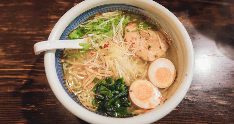 A steaming bowl of ramen topped with egg, pork and fresh greens sits on a wooden counter.
