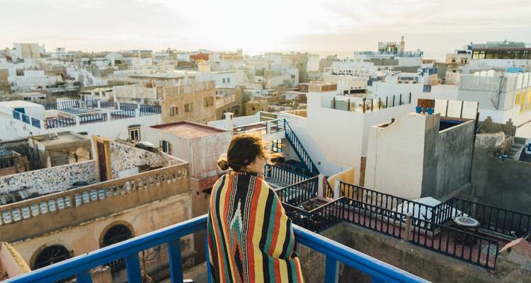 Gewikkeld in een kleurrijke deken, een vrouw kijkt uit over de witte daken van Essaouira vanaf een blauw balkon.