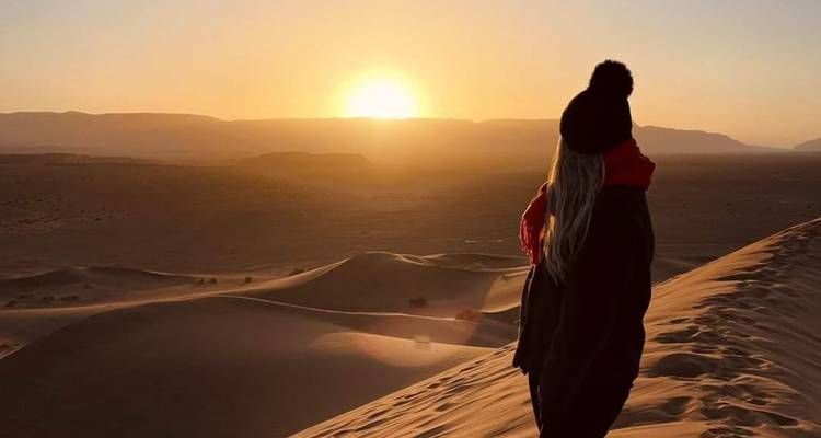 Een reiziger kijkt uit over golvende zandduinen naar een gloeiende zonsondergang in de Sahara.
