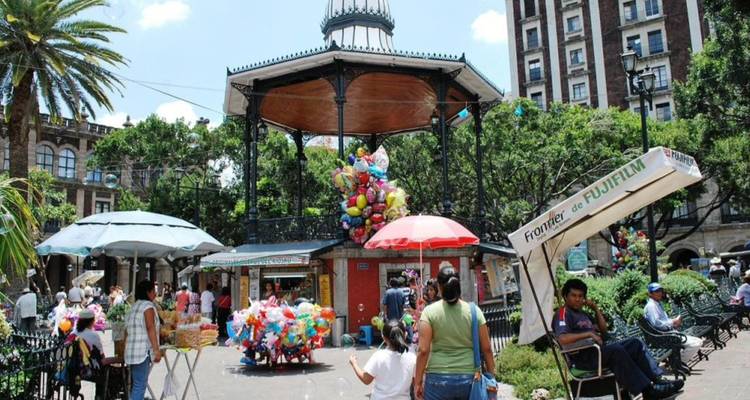 Plaza animada con quiosco, vendedor de globos y familias disfrutando de una tarde soleada entre palmeras.