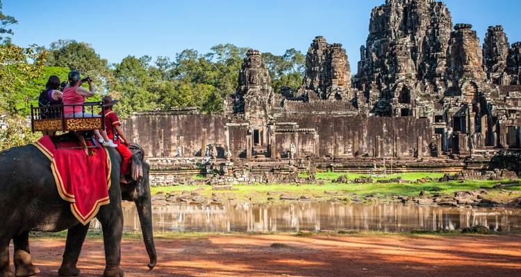 Une famille à cheval sur un éléphant passant devant les anciennes tours de pierre du temple de Bayon près d'une douve réfléchissante.