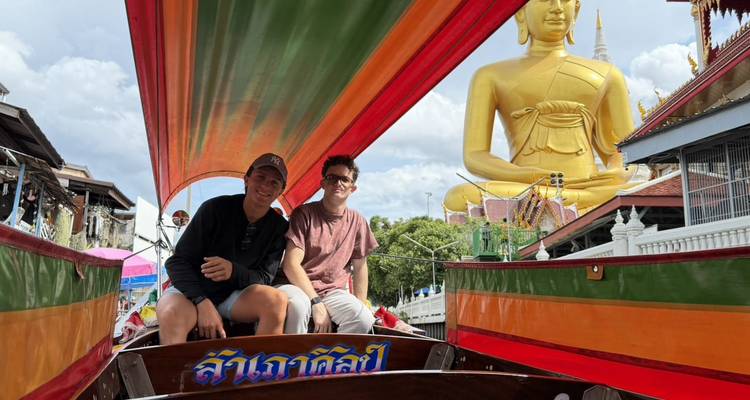 Two young travelers sit in a brightly coloured long-tail boat with a massive seated golden Buddha statue and temple buildings behind them.