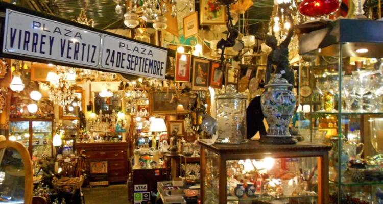 A cluttered antique shop filled with vintage furniture, lamps and curios, with old street signs hanging overhead.