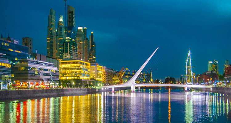 Night view of a modern city waterfront with the white Puente de la Mujer bridge reflected in the still water and illuminated skyscrapers behind.