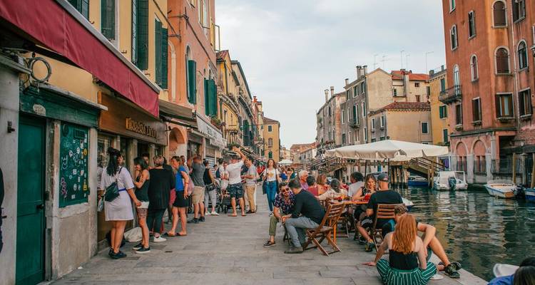 Crowds enjoy drinks along a lively canal-side promenade lined with colorful Venetian buildings.