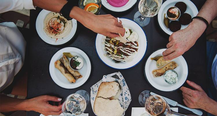 Overhead view of friends sharing assorted Mediterranean dishes and wine on a black table.