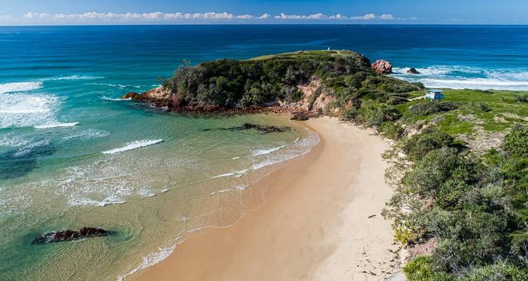 Vista aérea de una playa apartada en forma de media luna con agua turquesa cristalina y un promontorio cubierto de maleza.