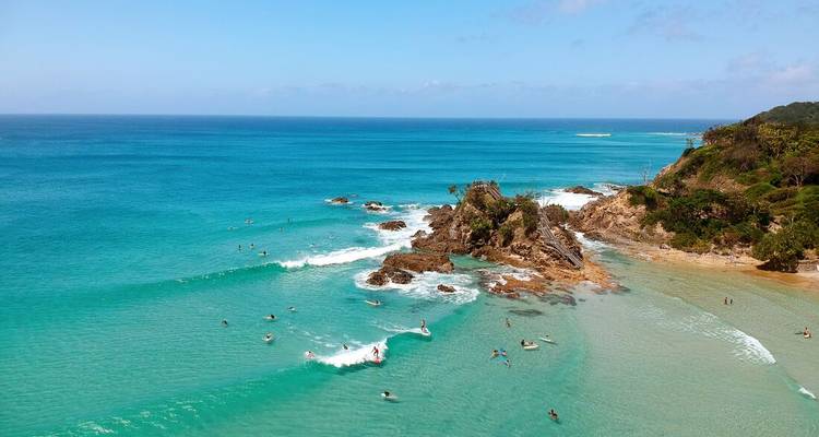 Surfers ride turquoise waves around rocky headland in crystal-clear ocean