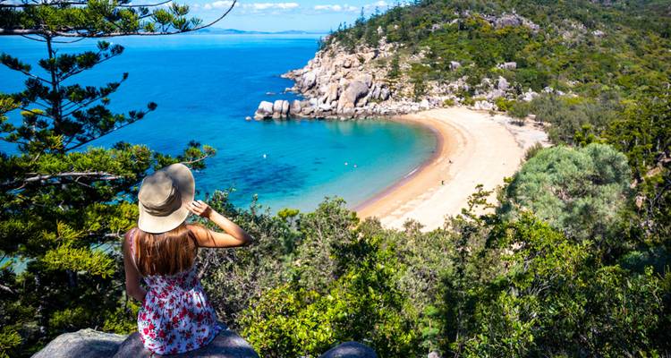 Woman in sunhat gazes over secluded bay with golden sand and turquoise water from cliff