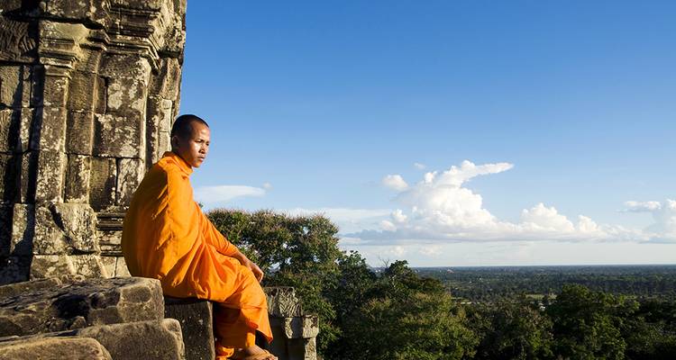 An orange-robed monk contemplates the vast Cambodian plain from an ancient sandstone temple ledge.
