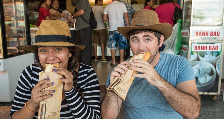 Travelers in hats happily bite into fresh bánh mì at a busy Vietnamese street stall.