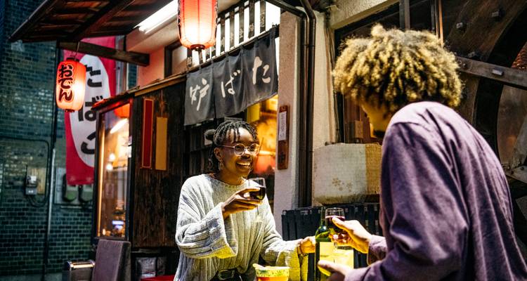 Deux amis partagent des boissons dans un izakaya intime éclairé par des lanternes sur une petite rue de Tokyo la nuit.