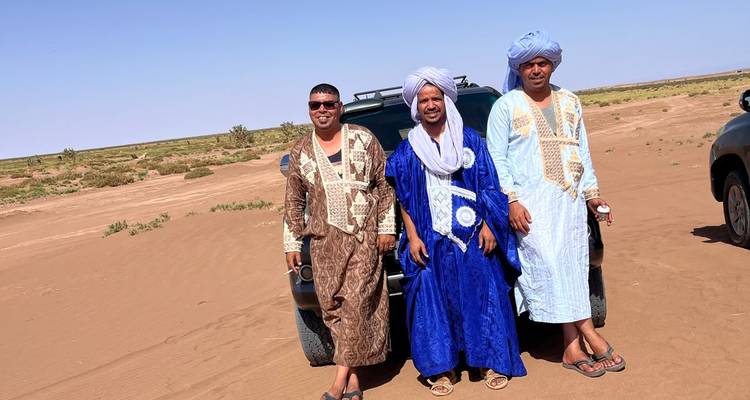 Trois hommes en robes traditionnelles posant près d'un véhicule utilitaire sport sur une plaine sableuse sous un ciel dégagé