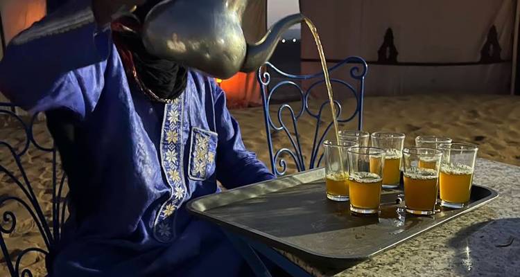 L'hôte berbère en robe bleue verse le thé à la menthe dans des verres sur un plateau à l'intérieur du camp du désert.