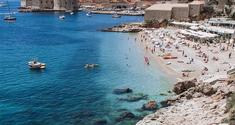 Busy city beach with sunbathers and kayaks beside medieval walls and turquoise Adriatic Sea