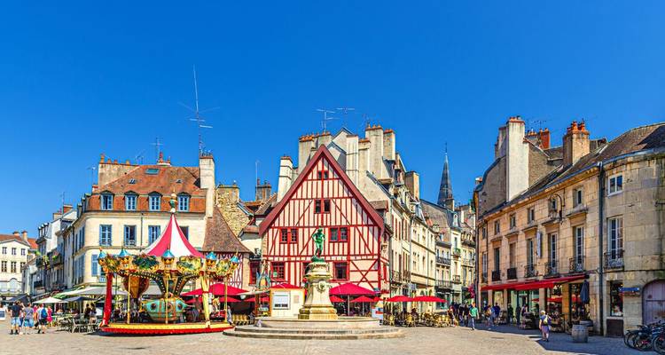 Maisons à colombages colorées, carrousel et fontaine dans une place française animée sous un ciel dégagé