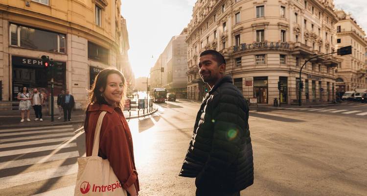 Deux voyageurs souriants traversant une rue ensoleillée bordée de bâtiments historiques élégants à Rome.