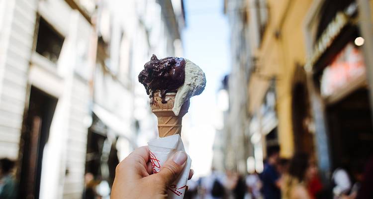 Gros plan d'une main tenant un cornet de glace avec une rue italienne floue et des bâtiments en arrière-plan.