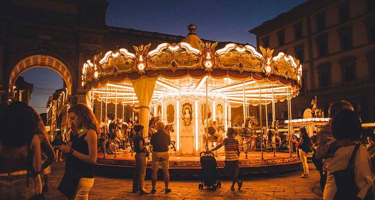 Carrousel vintage illuminé tournoyant sur une place italienne animée la nuit avec des personnes rassemblées autour.
