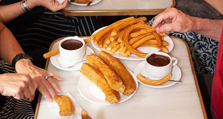 Des assiettes de churros avec des tasses de chocolat chaud épais sont partagées à une table de café tandis que des mains s'en saisissent.