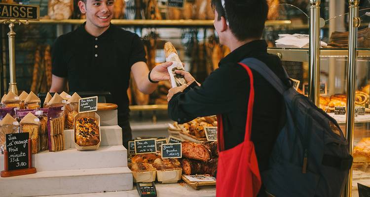Un boulanger bienveillant remet une baguette à un client à l'intérieur d'une boulangerie française remplie de pâtisseries.