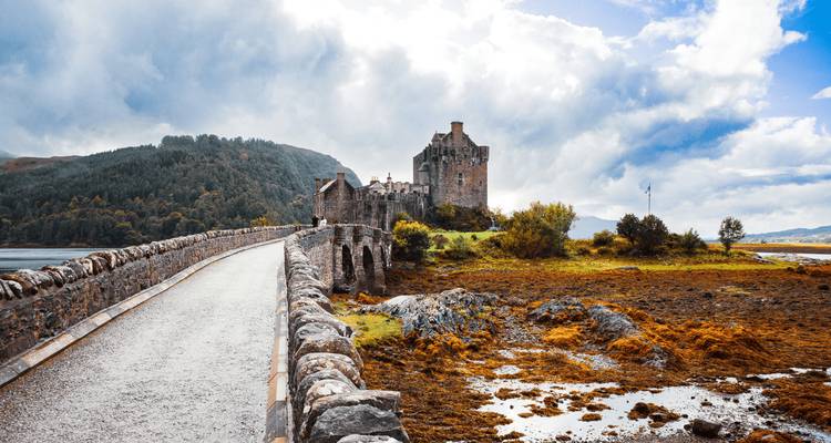 Stone bridge leading to a medieval castle set beside a loch with dramatic clouds and forested hills in the Highlands.
