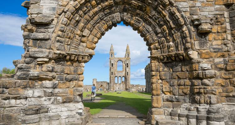 View through a grand stone archway of ancient cathedral ruins with a blue sky and green lawns beyond.