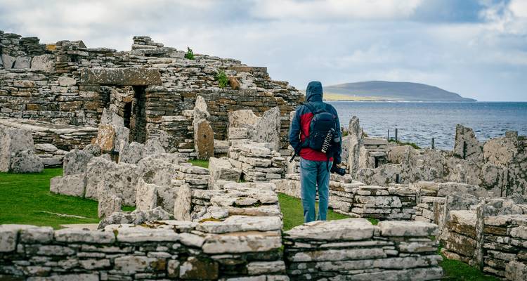 Hooded traveller standing among ancient stone ruins on a grassy cliff overlooking the sea and distant island.