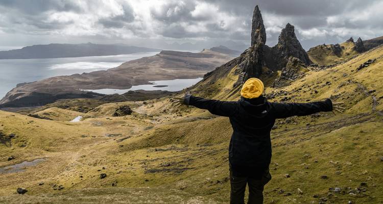 Traveller with outstretched arms atop a rugged ridge facing the dramatic pinnacles of the Old Man of Storr and sweeping sea views.