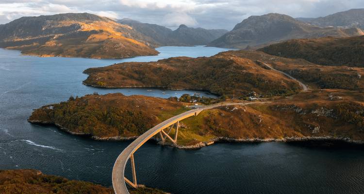 Dramatic aerial view of a sweeping bridge curving between rugged peninsulas over deep blue fjord-like waters.