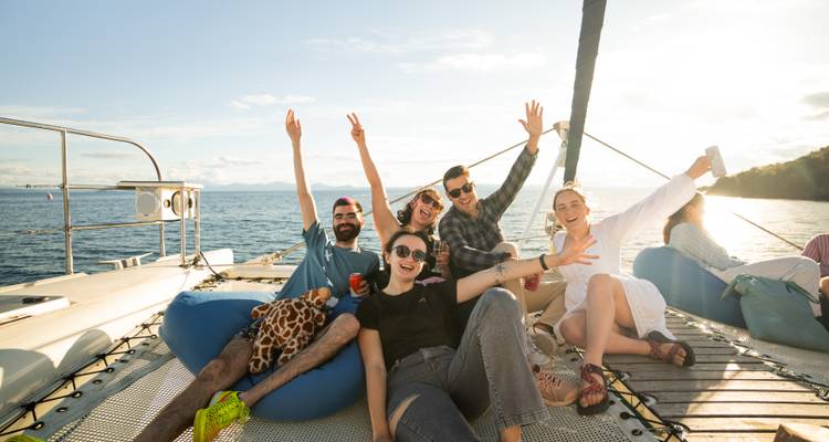 Group of friends celebrating on catamaran deck with ocean and sunset behind.