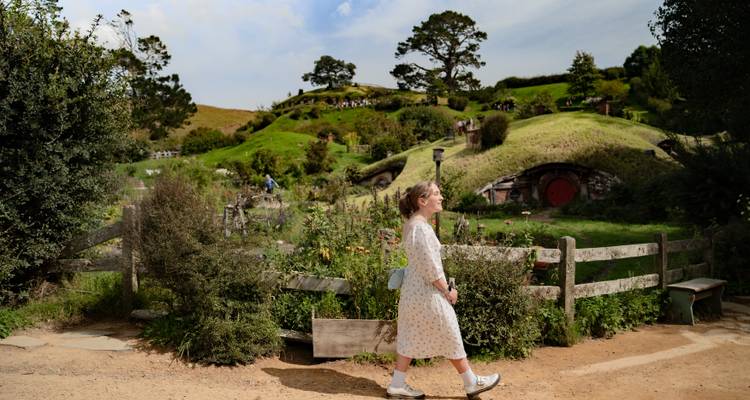 Young woman walking past the grassy hobbit-hole houses and gardens of the Hobbiton Movie Set in rolling green countryside under partly cloudy skies.