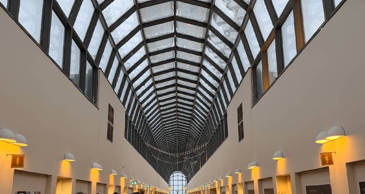 Corridor avec atrium aux parois de verre, poutres symétriques et appliques murales chaleureuses.
