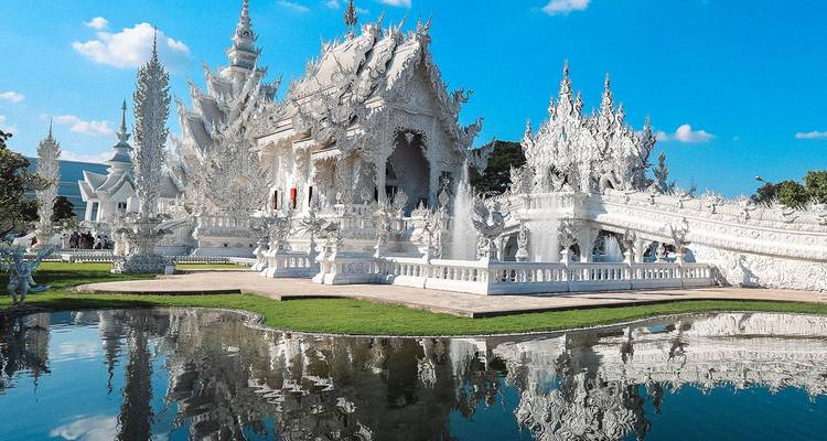 Dazzling all-white Wat Rong Khun temple reflected perfectly in a still pond under a bright sky.