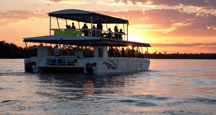A river cruise boat filled with silhouetted passengers glides over calm water at a fiery sunset.