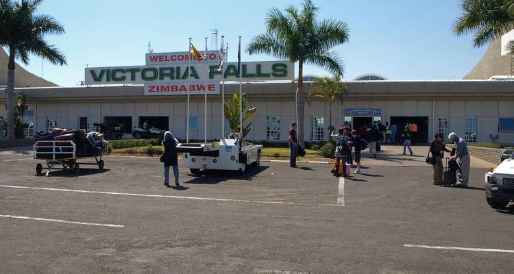 Travellers with luggage outside Victoria Falls Airport under a large welcome sign.