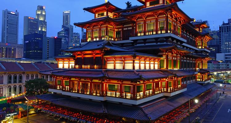 The richly illuminated Buddha Tooth Relic Temple glowing against the evening sky with modern skyscrapers behind