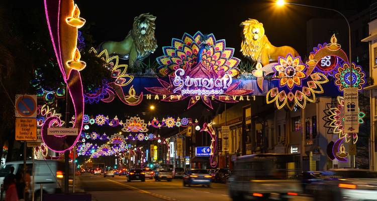 Bustling street decorated with vibrant Deepavali light arches and lion figures, cars and pedestrians passing underneath at night
