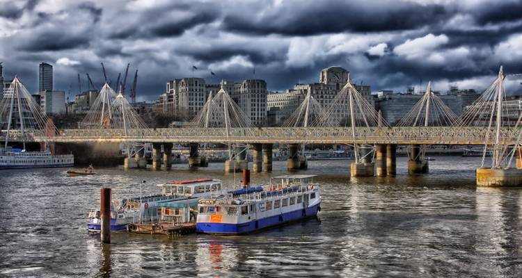 Moody view of boats docked on a wide river with dramatic storm clouds over a suspension footbridge and city skyline