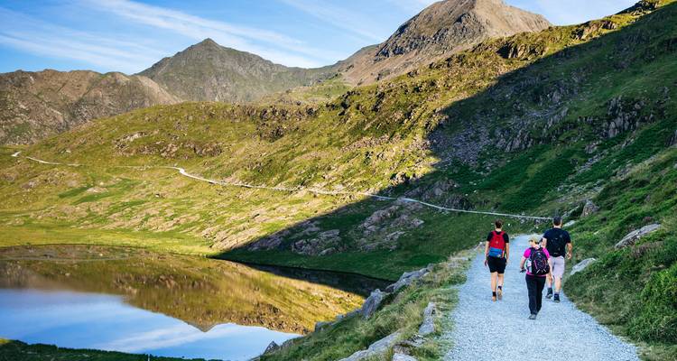 Hikers follow a gravel path beside a reflective alpine lake beneath rugged peaks in Snowdonia.