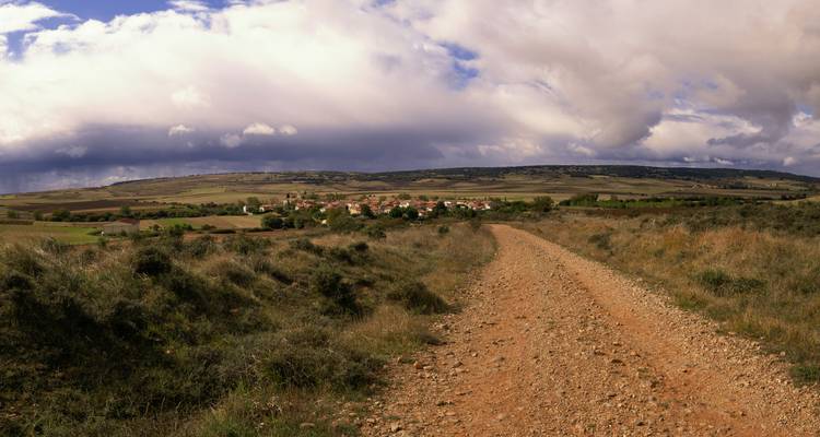 Wide gravel path leading to a small Spanish village under dramatic cloudy sky
