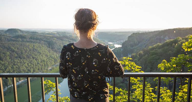 Femme se tenant à une balustrade surplombant une vallée verdoyante luxuriante et une rivière sinueuse au coucher du soleil.
