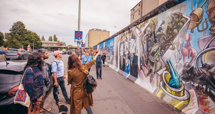 Un guide touristique fait des gestes en direction d'une fresque murale colorée de la East Side Gallery de Berlin tandis qu'un petit groupe écoute.